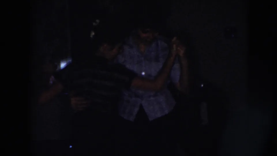 Two adults dance closely in a dim Nebraska home interior, with faces and clothing only partly visible in the low light.