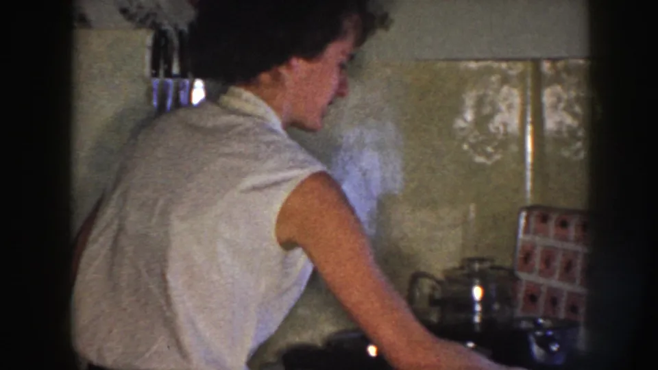 A woman in a sleeveless light blouse works at a stove in a tiled Nebraska kitchen, with pots and a kettle nearby.