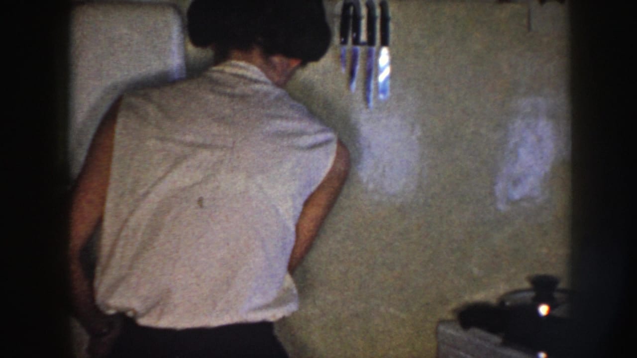 In a Nebraska 1958 home-movie frame, a woman in a sleeveless white blouse stands with her back to the camera in a kitchen.