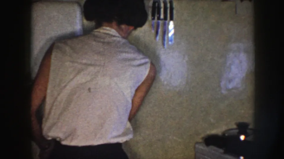 In a Nebraska 1958 home-movie frame, a woman in a sleeveless white blouse stands with her back to the camera in a kitchen.