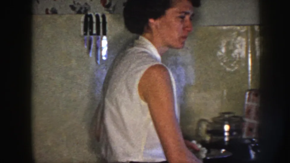 The woman turns in profile while working beside cookware in a modest 1958 Nebraska kitchen interior.