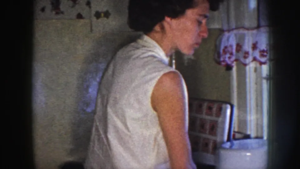 The woman stands near a kitchen window with floral curtains, shown in grainy 8mm home-movie footage from Nebraska in 1958.