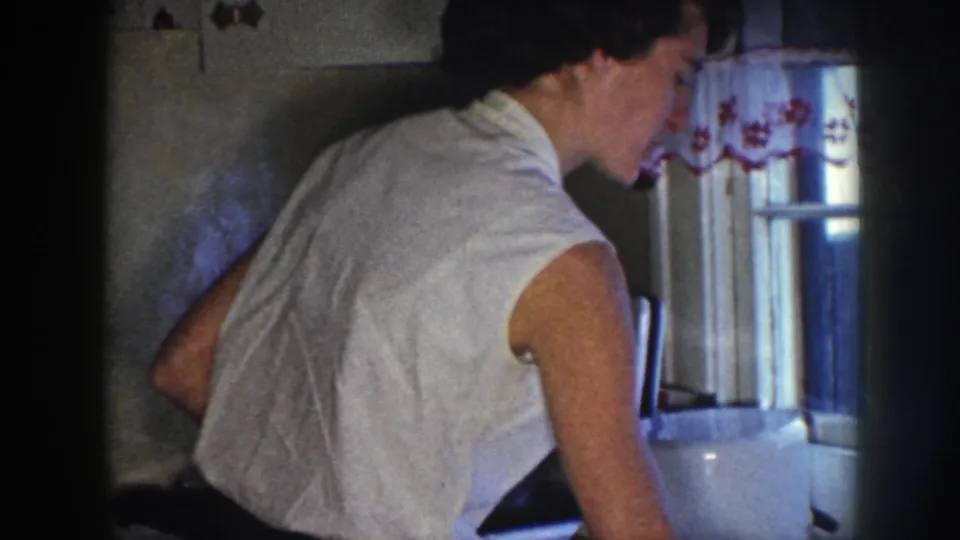 A woman leans near the counter by a curtained window in a private Nebraska kitchen, captured on 8mm film in 1958.