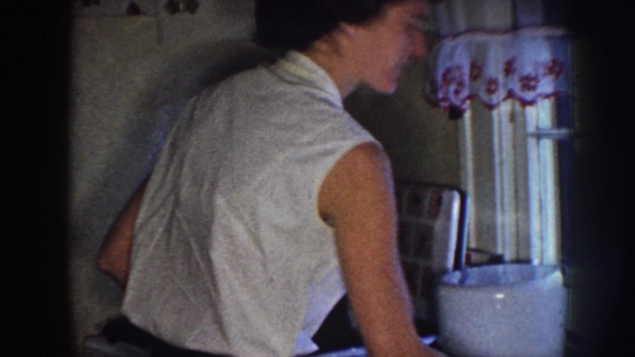 Woman in a white sleeveless dress stands at a dim kitchen counter beside a curtained window.