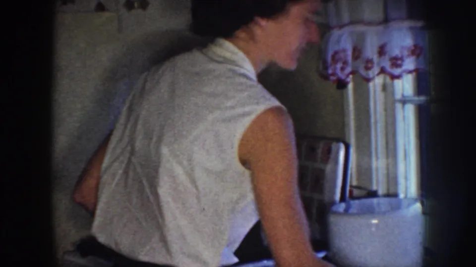 In a 1958 Nebraska home-movie frame, a woman faces away in a dim kitchen near a floral curtain and countertop.
