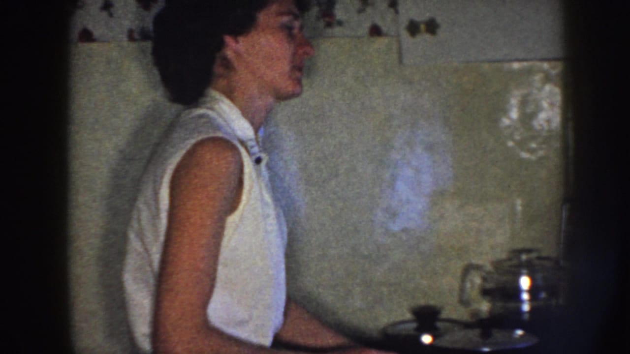 Woman in profile stands at a stove area with a metal teapot on the right.