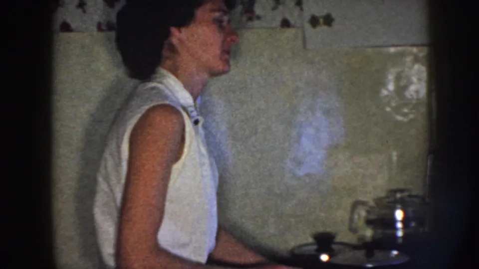 The woman appears in profile in a Nebraska kitchen, standing near a stovetop with a shiny teapot and tiled wall.