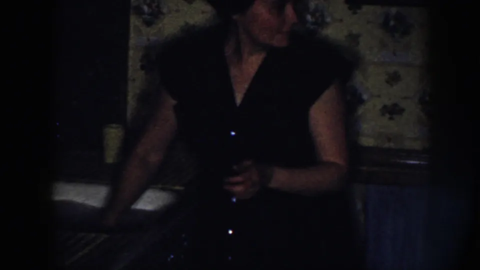 In 1958 Nebraska home-movie footage, a woman in a dark dress stands at a sink near floral wallpaper in a low-lit kitchen.