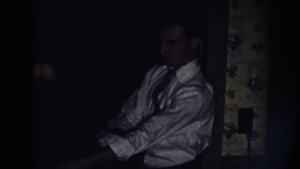 A man in a white button-up shirt and tie adjusts his sleeve in a shadowy 1958 Nebraska household interior.