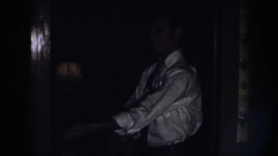 The man appears in profile wearing a white shirt and tie, partly silhouetted inside the dim Nebraska home interior.