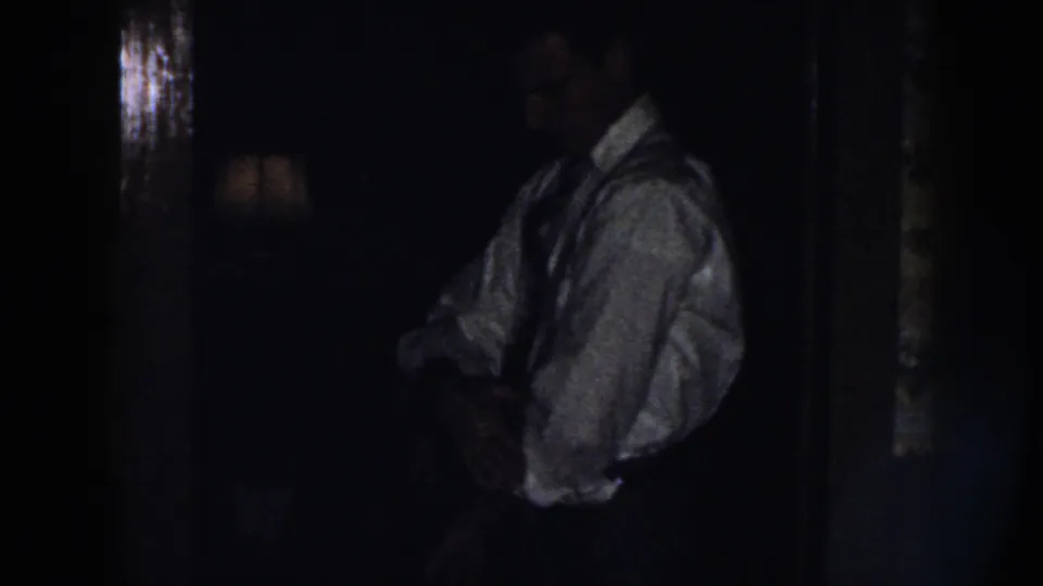A man in a white shirt and tie adjusts his clothing beside floral wallpaper in a low-light 1958 Nebraska home scene.