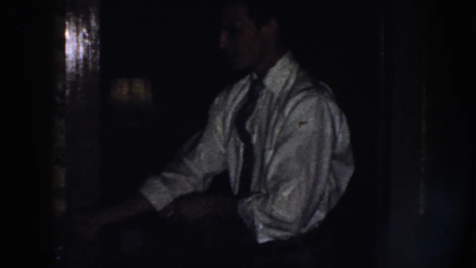 In a 1958 Nebraska home-movie frame, a man in a white shirt and tie stands in a dim kitchen near a dark surface.