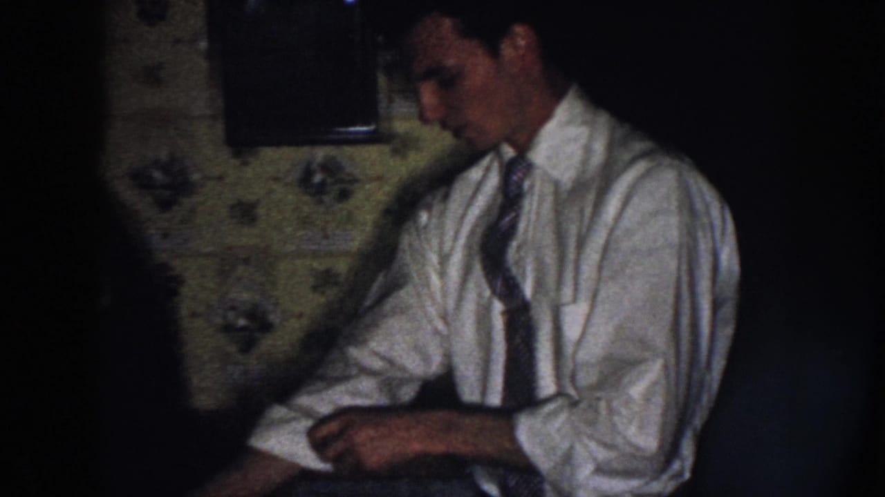 A man in a white shirt and tie appears to roll or adjust his sleeve beside floral wallpaper in a Nebraska kitchen scene.