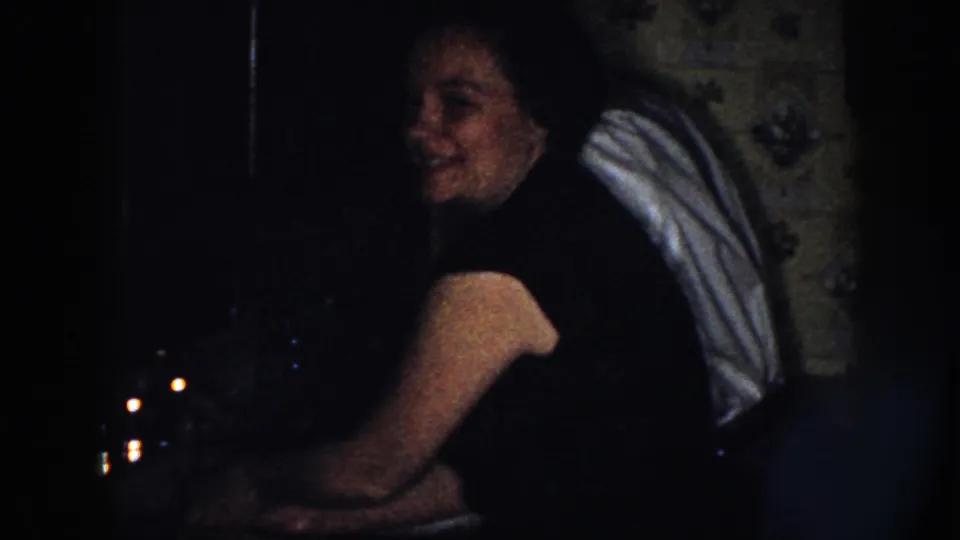 The woman turns from the sink and smiles faintly in a dim 1958 Nebraska kitchen or wash area.