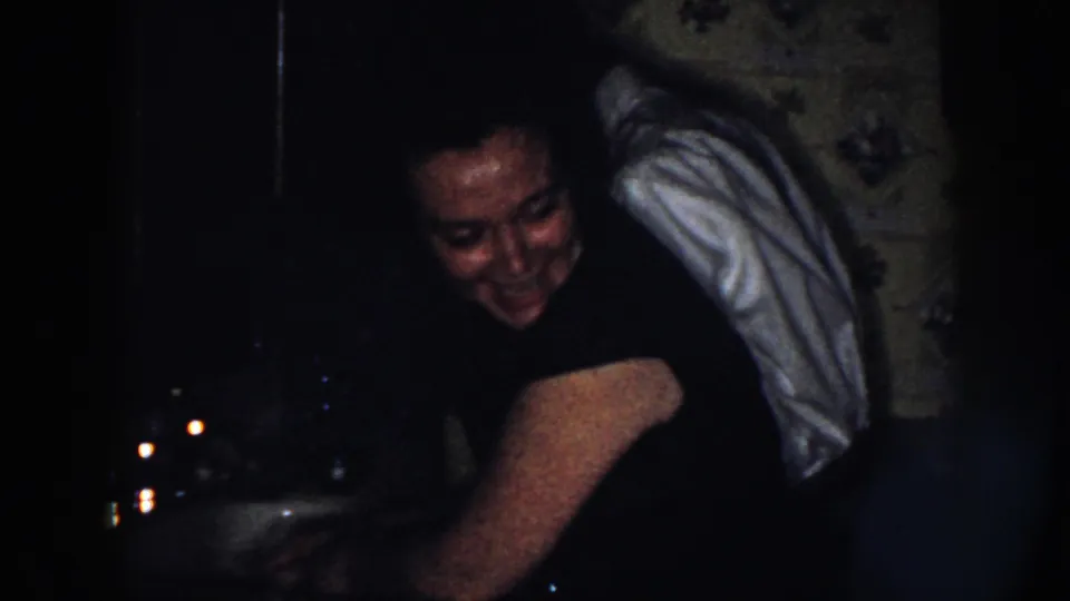 A woman leans over a kitchen sink in dim 1958 Nebraska home-movie footage, with her face partly lit.