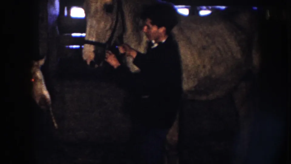 An adult man holds or adjusts the larger horse in the dim Nebraska stable, with another person partly visible nearby in the 1958 frame.