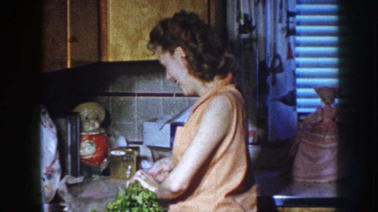 Woman in a 1950s kitchen prepares leafy vegetables at a counter, with cabinets and ceramic decor behind her.