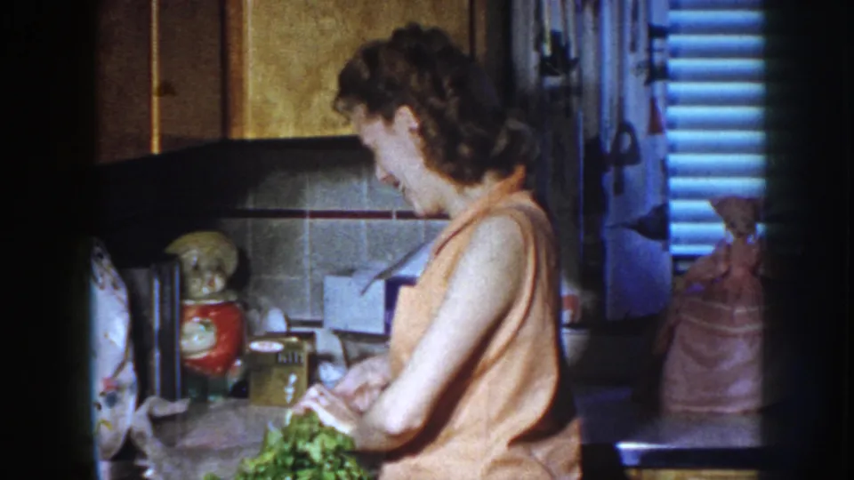 Woman in a 1950s kitchen prepares leafy vegetables at a counter, with cabinets and ceramic decor behind her.