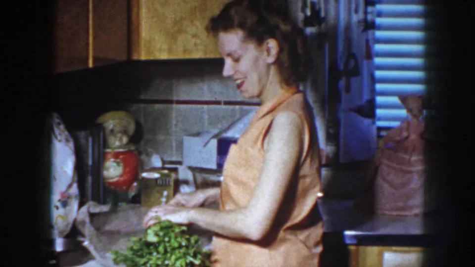 In the Nebraska 1958 home-movie scene, the woman smiles while preparing vegetables in a private kitchen.