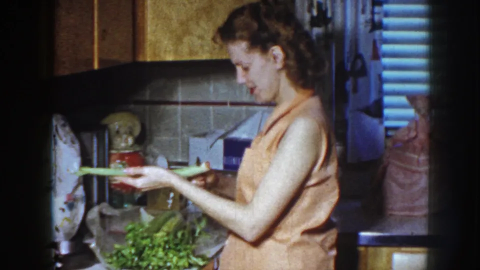 A 1958 home-movie frame shows the woman examining or trimming a long celery stalk over a pile of greens in the kitchen.