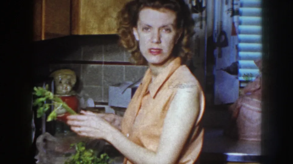 The woman turns toward the camera while holding greens in the 1958 Nebraska kitchen, creating a candid home-movie moment.