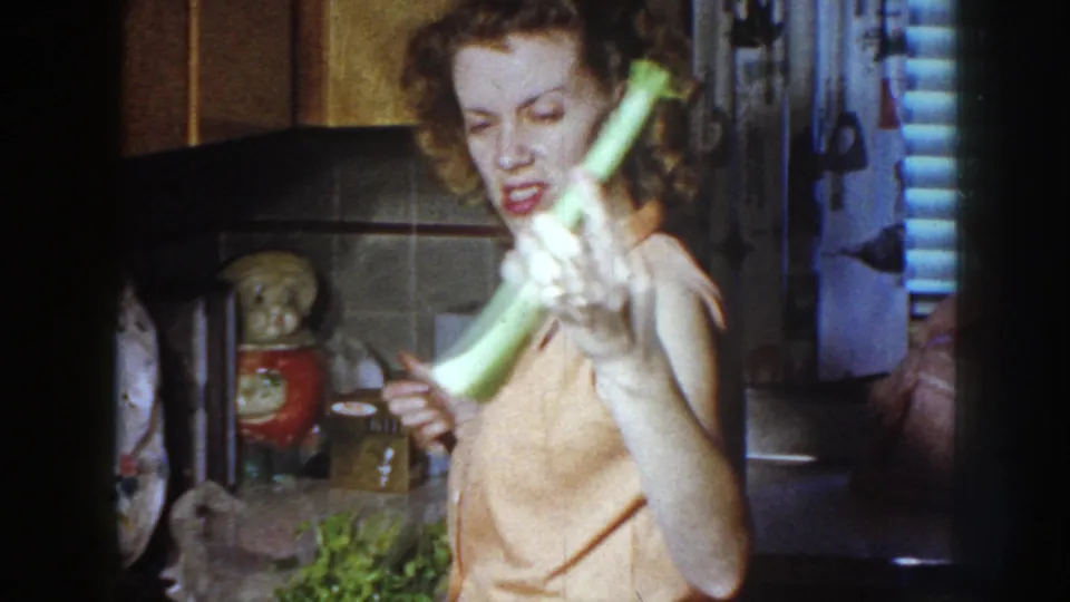 A woman in a 1958 Nebraska kitchen faces the camera, holding celery forward in one hand and a paring knife in the other.
