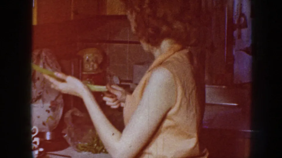 The woman is viewed from behind in the kitchen, holding celery and a paring knife as warm faded film color shifts across the frame.