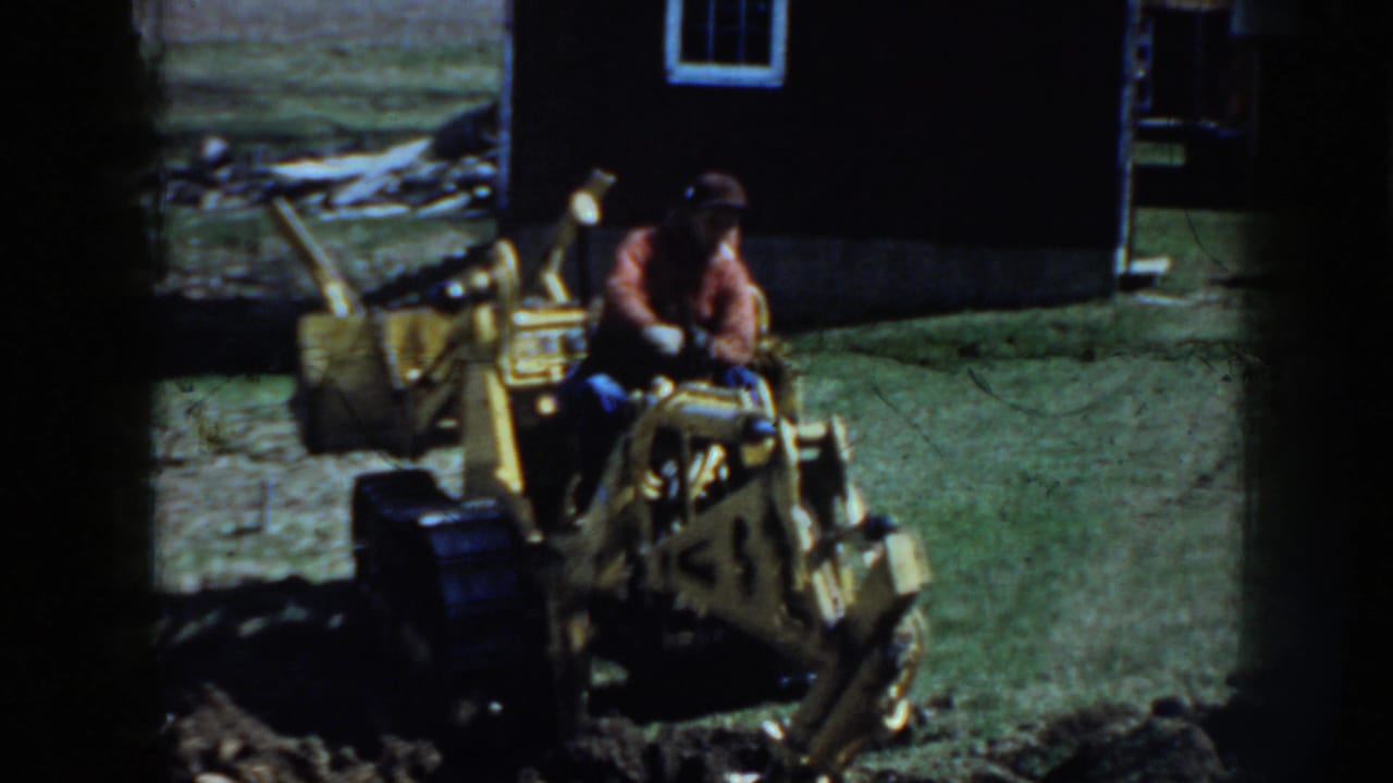In 1958 Nebraska home-movie footage, a man in a red shirt and cap operates a yellow tracked backhoe beside a dark barn-like building.