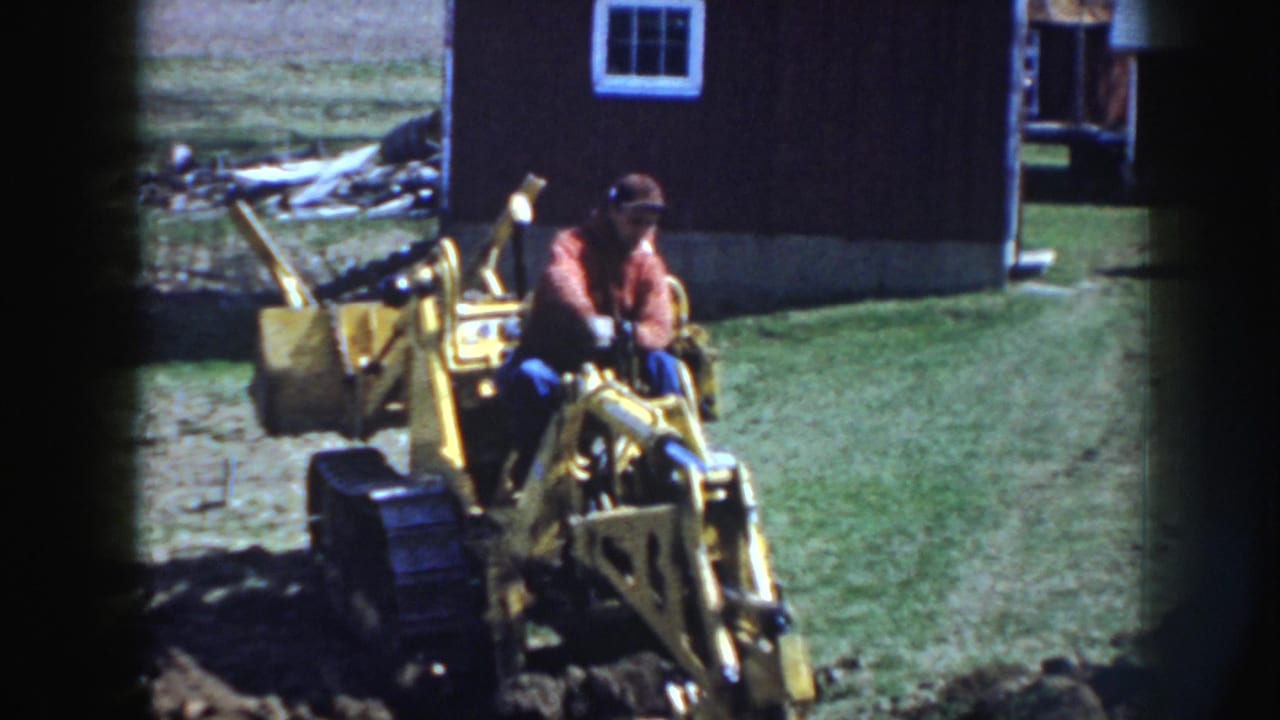 The operator sits forward on the yellow backhoe as it moves over grass and soil near a dark rural outbuilding in Nebraska.
