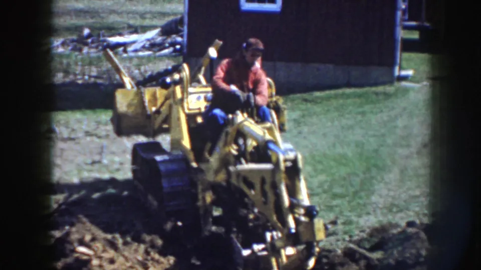 A man in work clothes controls the yellow tracked backhoe, with churned soil in the foreground and a barn-like structure behind him.