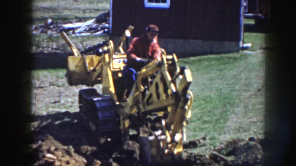 The yellow backhoe's arm and bucket appear in front as the seated operator works near grass, soil, and a dark rural building.