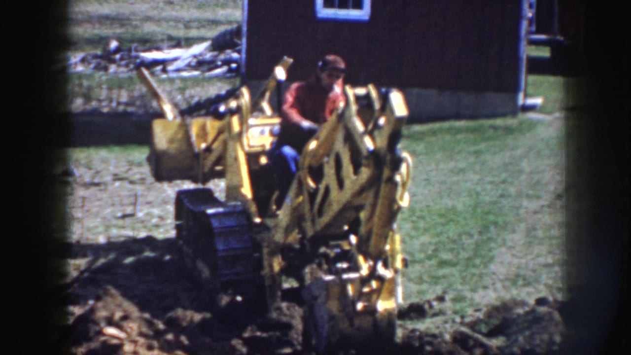 Man in a red shirt operates a yellow backhoe beside a dark wooden building.