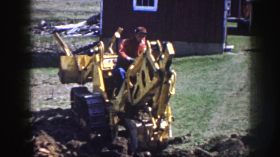 Home-movie frame of a man operating a yellow backhoe beside a dark wooden building in Nebraska, 1958.