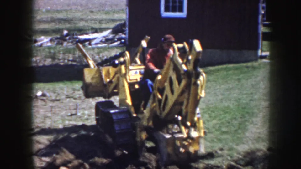 A 1958 Nebraska 8mm frame shows a man in red operating a yellow backhoe near exposed soil and a wooden outbuilding.