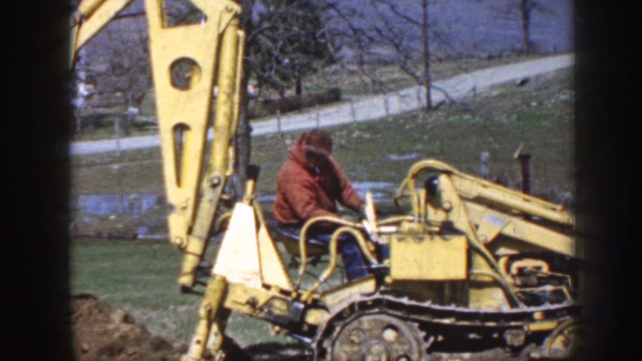 Man in a red jacket operates a yellow tracked backhoe on grassy ground.