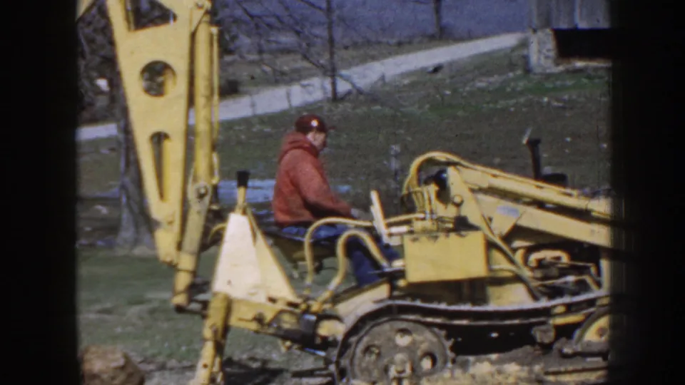 A 1958 Nebraska frame shows the man seated on the yellow tracked backhoe as the boom and stabilizer area fill the foreground.