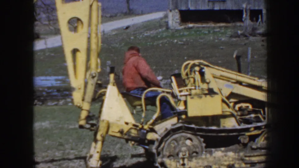 The final 1958 Nebraska frame shows the operator from behind on the yellow backhoe, with grass, mud, a road, and bare trees nearby.