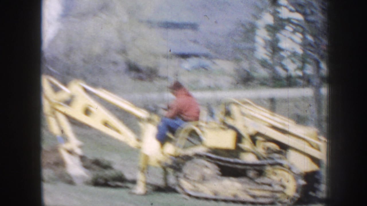 Man on a yellow tracked backhoe digging in a grassy yard.