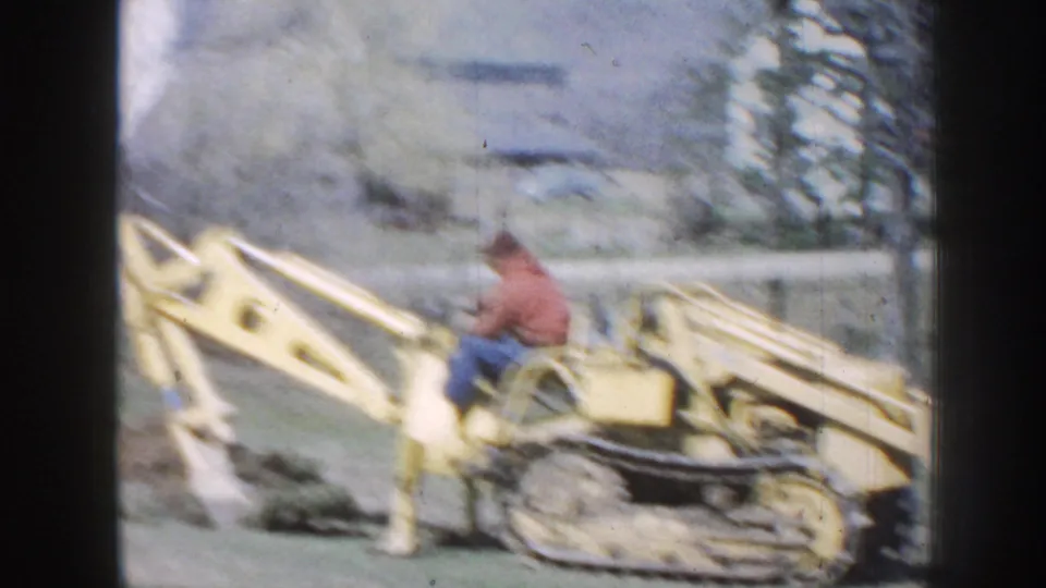 Soft 8mm frame of a man operating a yellow tracked backhoe on a grassy Nebraska property in 1958.
