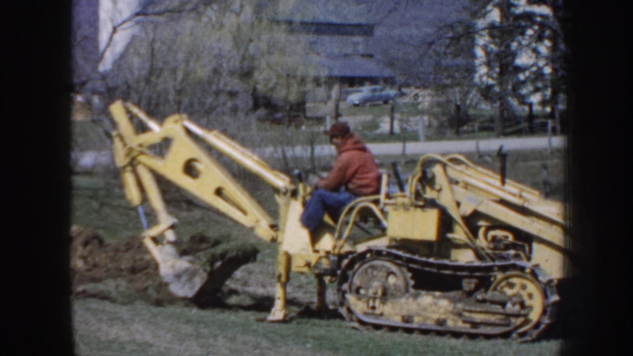 Yellow tracked backhoe with operator beside a dug area.