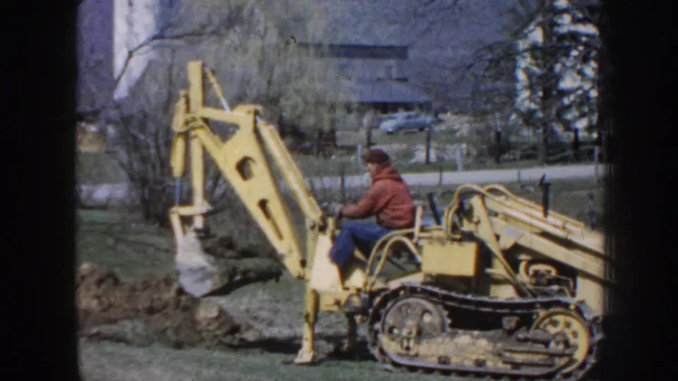 1958 Nebraska home-movie frame showing the backhoe bucket lowered near a hole while the seated operator watches the work area.