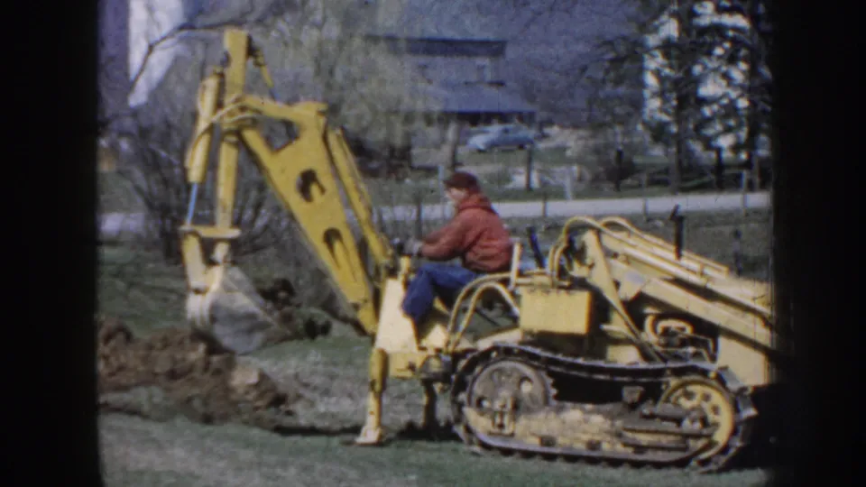 Man seated on a yellow tracked backhoe with its arm raised over disturbed soil on a rural Nebraska property in 1958.