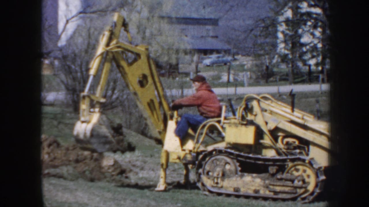Man in a red jacket operates a yellow tracked excavator beside a mound of earth.