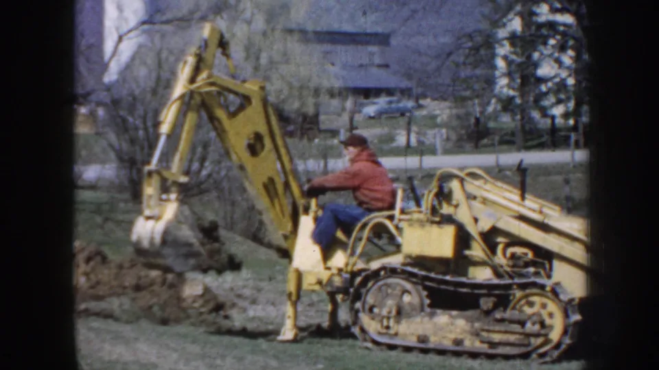In rural Nebraska, 1958, a man sits on a yellow tracked excavator as its bucket digs into earth on a grassy property.