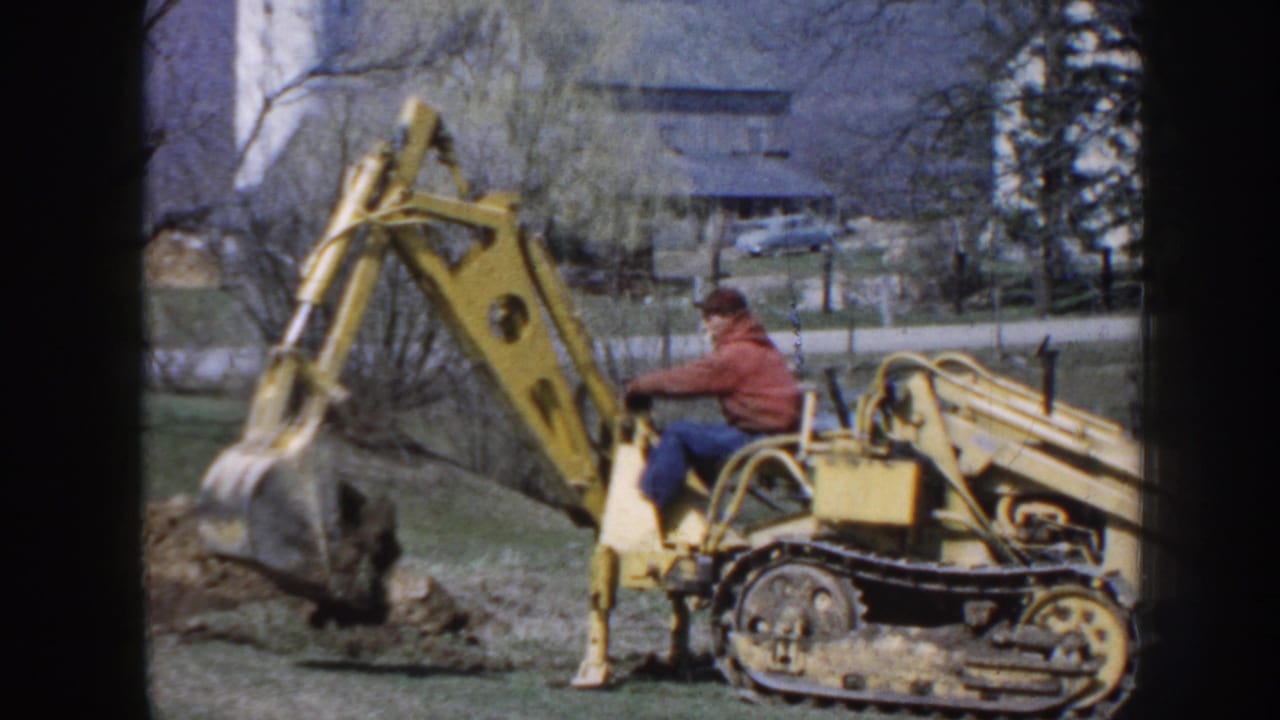 The 1958 Nebraska home-movie frame shows the operator guiding a yellow tracked excavator as the bucket lifts dirt.
