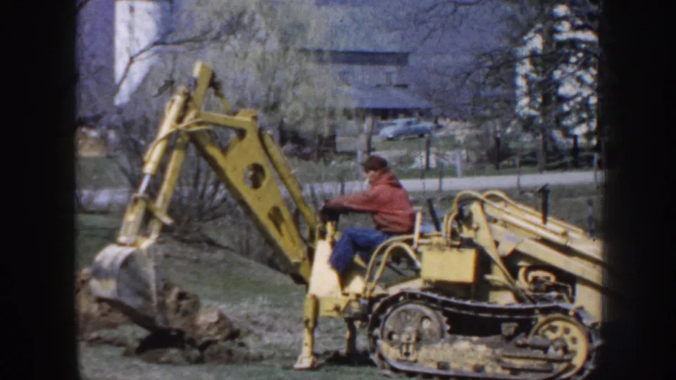 A man in work clothes operates a yellow tracked excavator on rural Nebraska property, with the bucket near the soil.