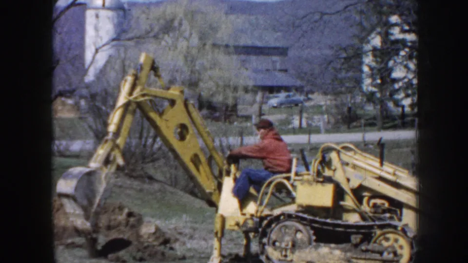 The operator remains on the yellow tracked excavator while the bucket shifts beside disturbed earth in a 1958 Nebraska rural scene.