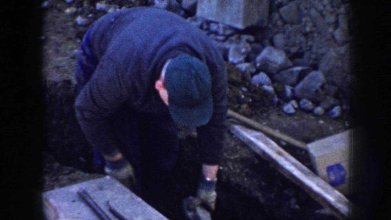 Man in a dark jacket and green cap bends over tools beside a trench and rocks.