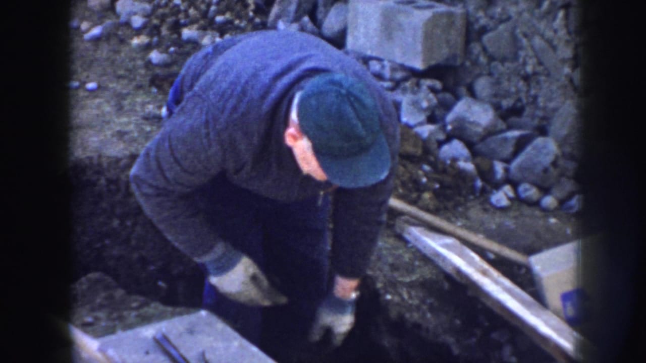 The worker crouches at the rail construction site, reaching toward the ground near rocks, boards, concrete, and a cardboard box.