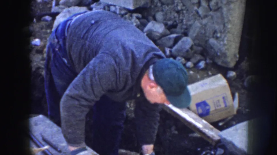In the 1958 Nebraska home-movie frame, the man leans forward over a rough work area with a box, plank, rocks, and dirt around him.